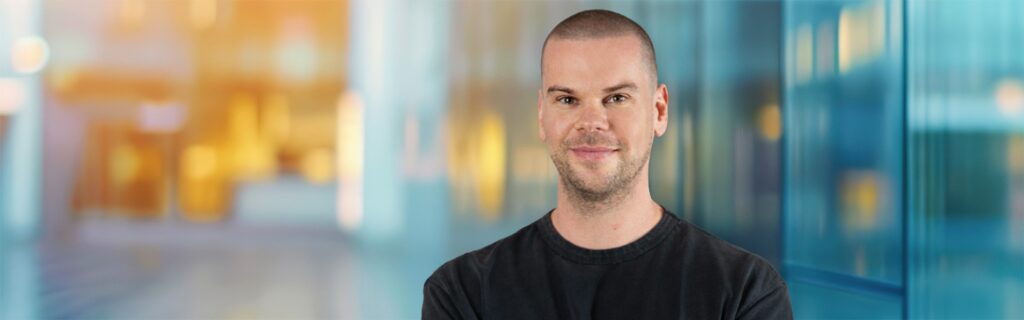 Ein Mann mit rasiertem Kopf und schwarzem T-Shirt steht lächelnd vor einem unscharfen, modernen Innenhintergrund mit blauen und gelben Farbtönen, während er über S&P 500 Optionen spricht.